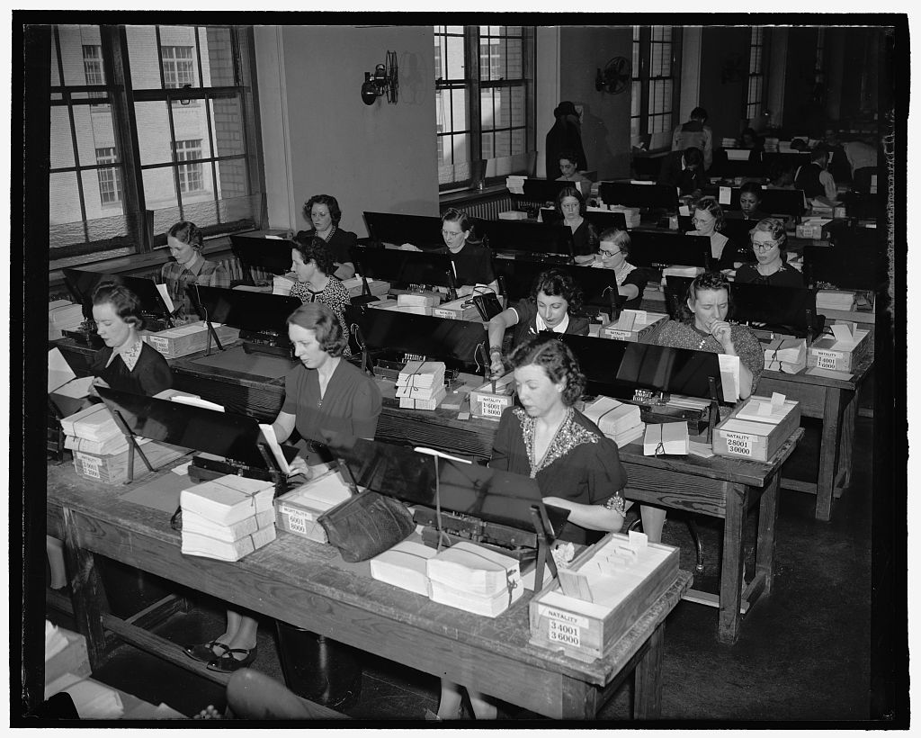 Women work at long rows of Census Bureau tabulating machines, surrounded by stacks of punched cards.