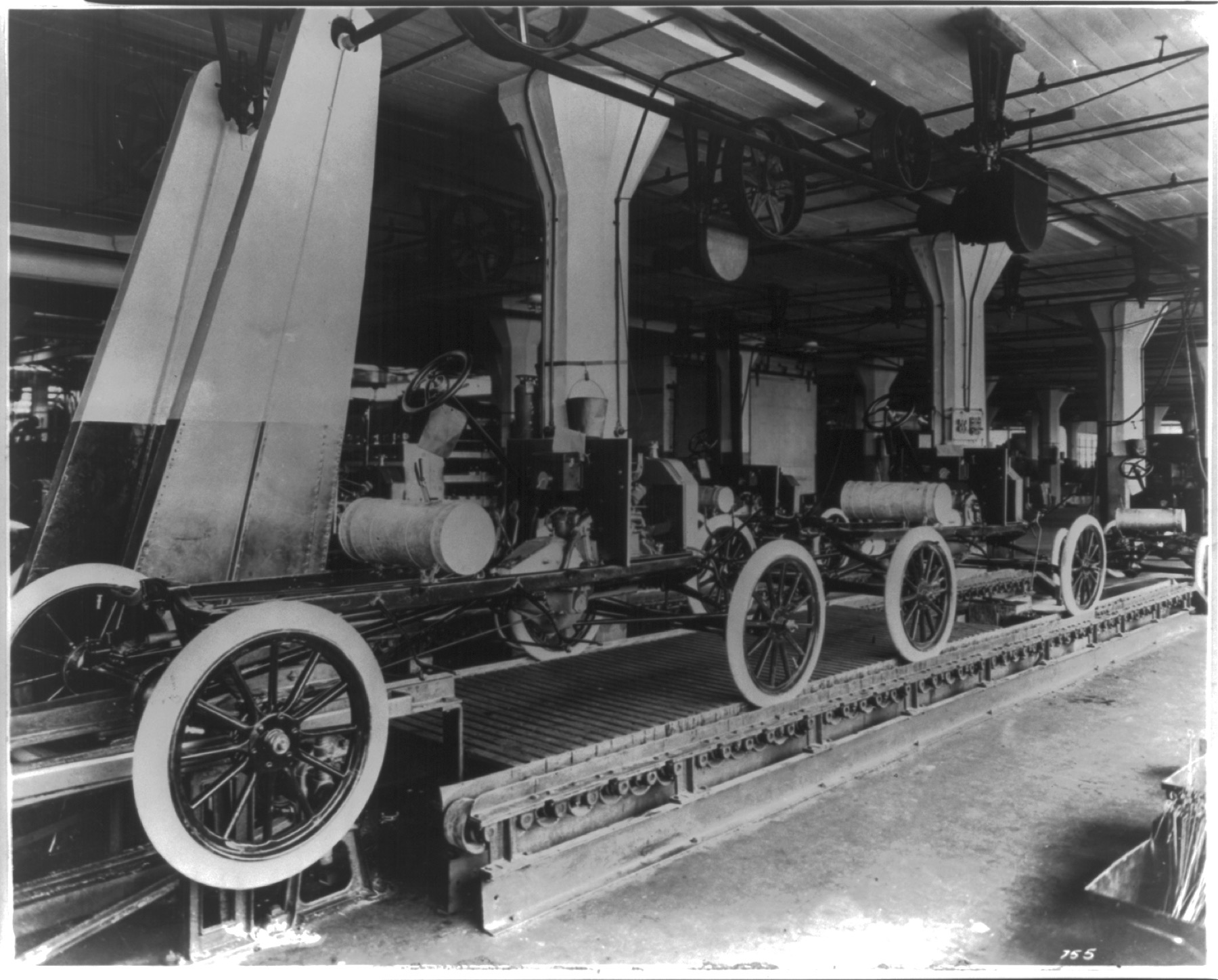 Automobile chassis move in a row beneath belts, wheels, and other machinery on an early Ford assembly line.