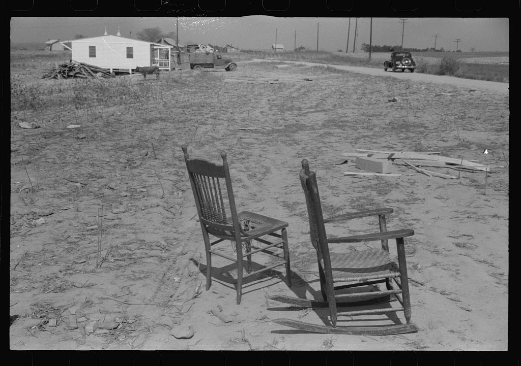 Two wooden chairs sit in a cleared field near a small prefabricated house, a truck, and a rural road.