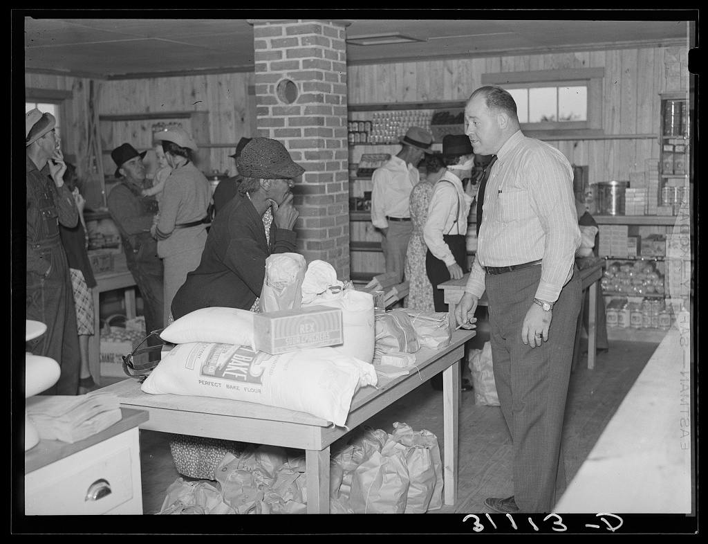 A customer and store worker speak across a table of flour, crackers, and other goods while people shop behind them.