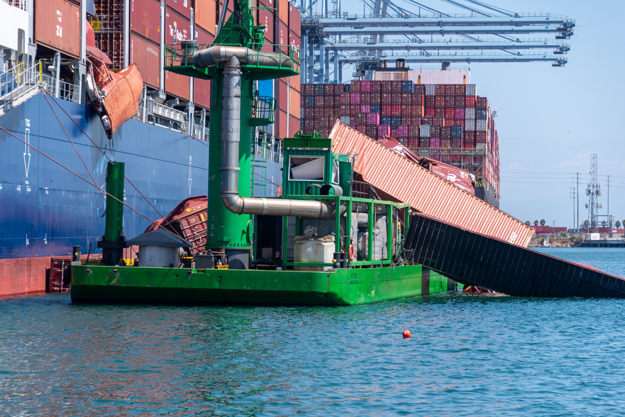 Fallen shipping containers lean from a cargo ship across a green emissions-control barge in the Port of Long Beach.