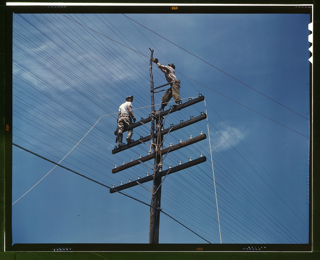 Two workers balance high on a wooden pole while maintaining a dense network of telephone lines.