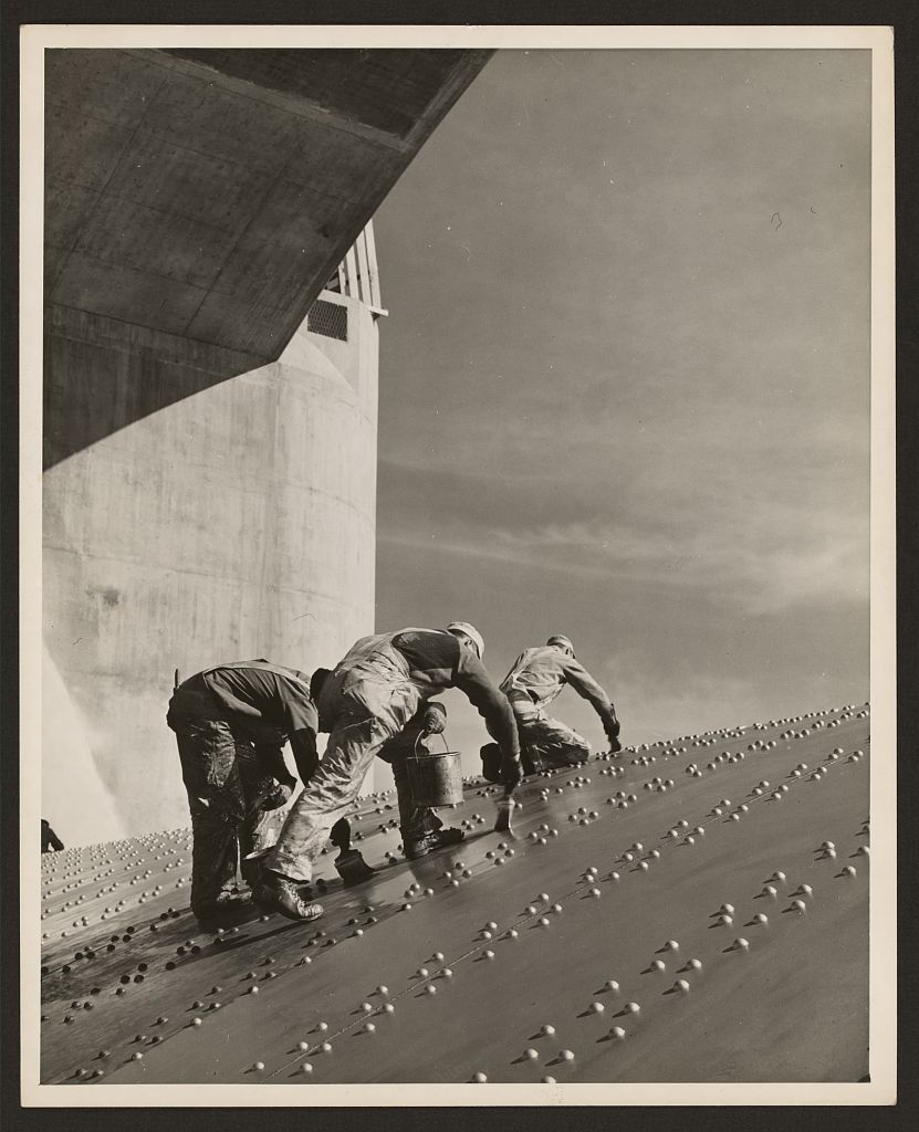 Three workers crouch on the steep riveted-steel wall of a Hoover Dam spillway while applying a protective coat.